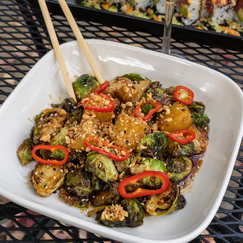 A white rectangular dish of J Liu's roasted Brussels sprouts with red peppers, peanuts, and sesame seeds.