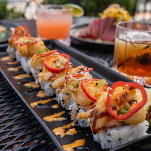 A long black plate of J Liu's Asian-fusion sushi rolls with drinks and other dishes in the background.